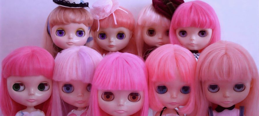 Nine large-eyed fashion dolls with pink and pastel hair arranged closely together in two rows against a soft pink background.